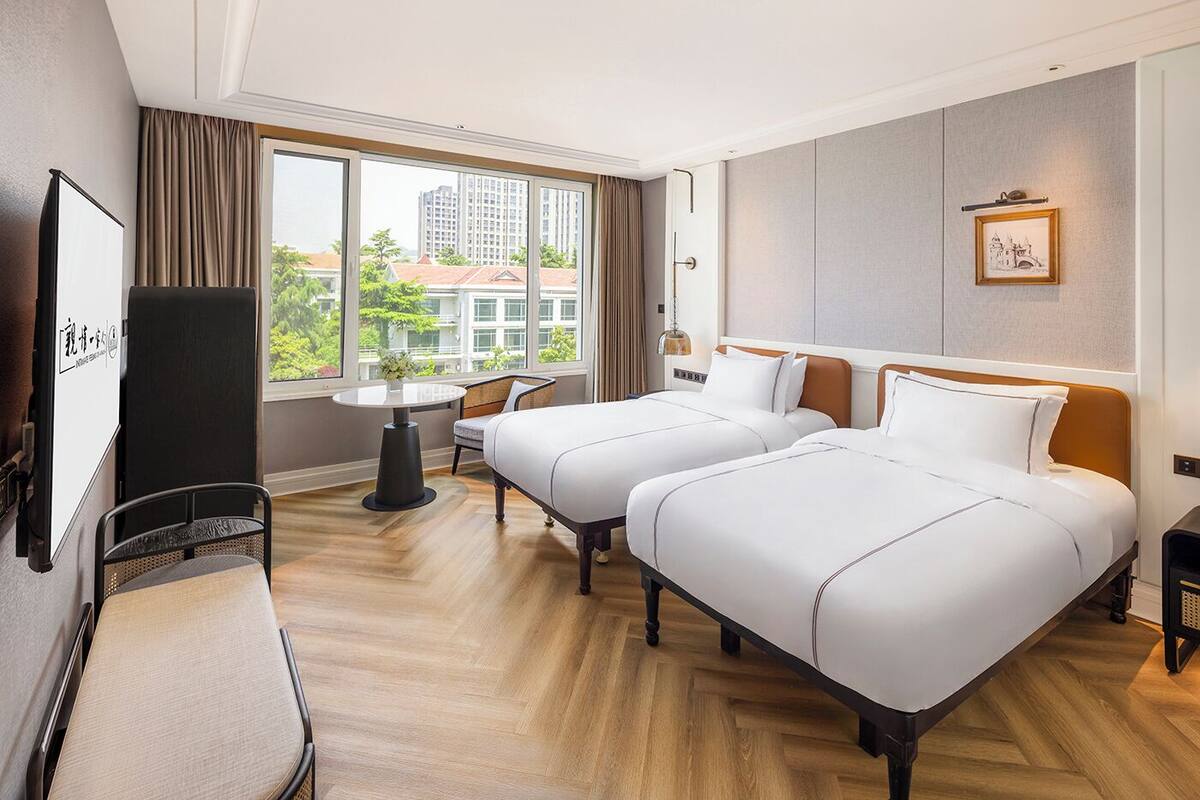 A hotel room with two beds, a desk, a chair, a TV, and a window with a city view.