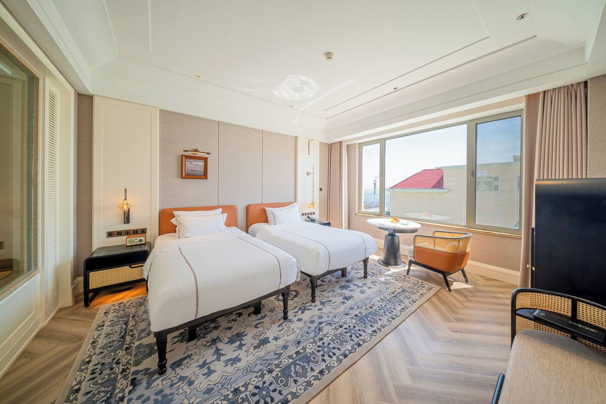 A hotel room with two beds, a TV, a small table, and a window with a view of buildings.