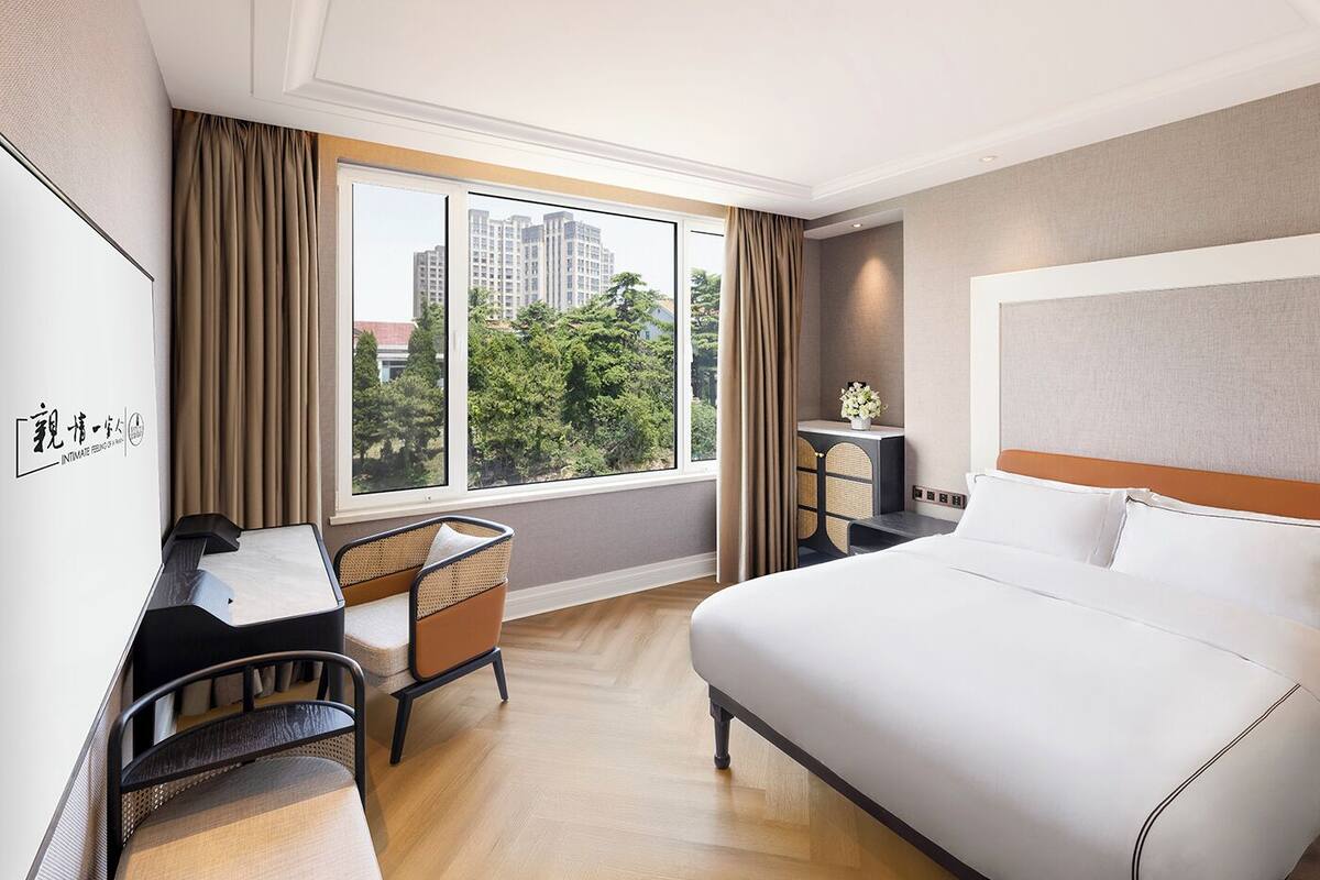 A hotel room with a bed, a desk with a chair, a small table with a vase of flowers, and a view of buildings and trees through the window.