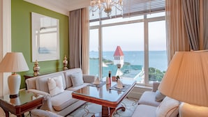 Ein Hotelzimmer mit großem Fenster, das Meer und einen Turm zeigt, einer Kristallleuchtre, einem Sofa und einem Tisch mit Dekorationen.