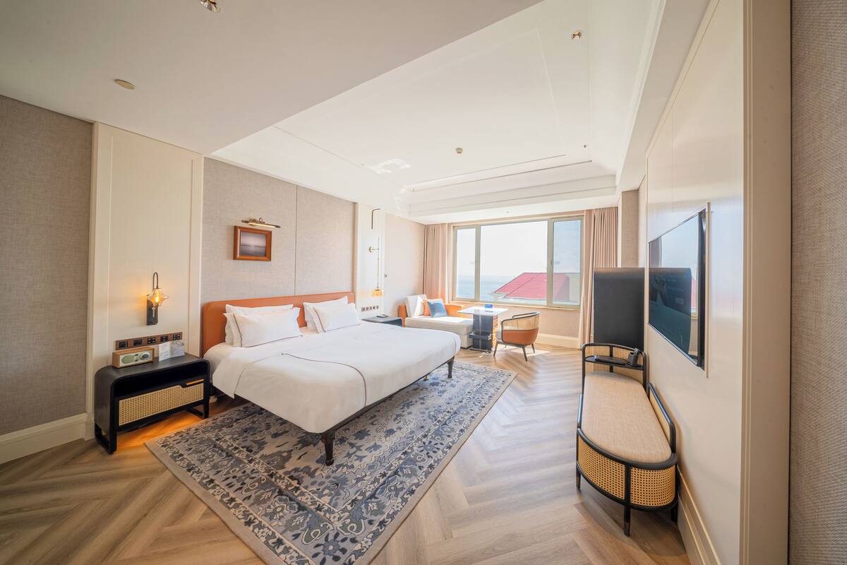A modern hotel room with a large bed, a desk, a chair, a TV, and a view of the city.