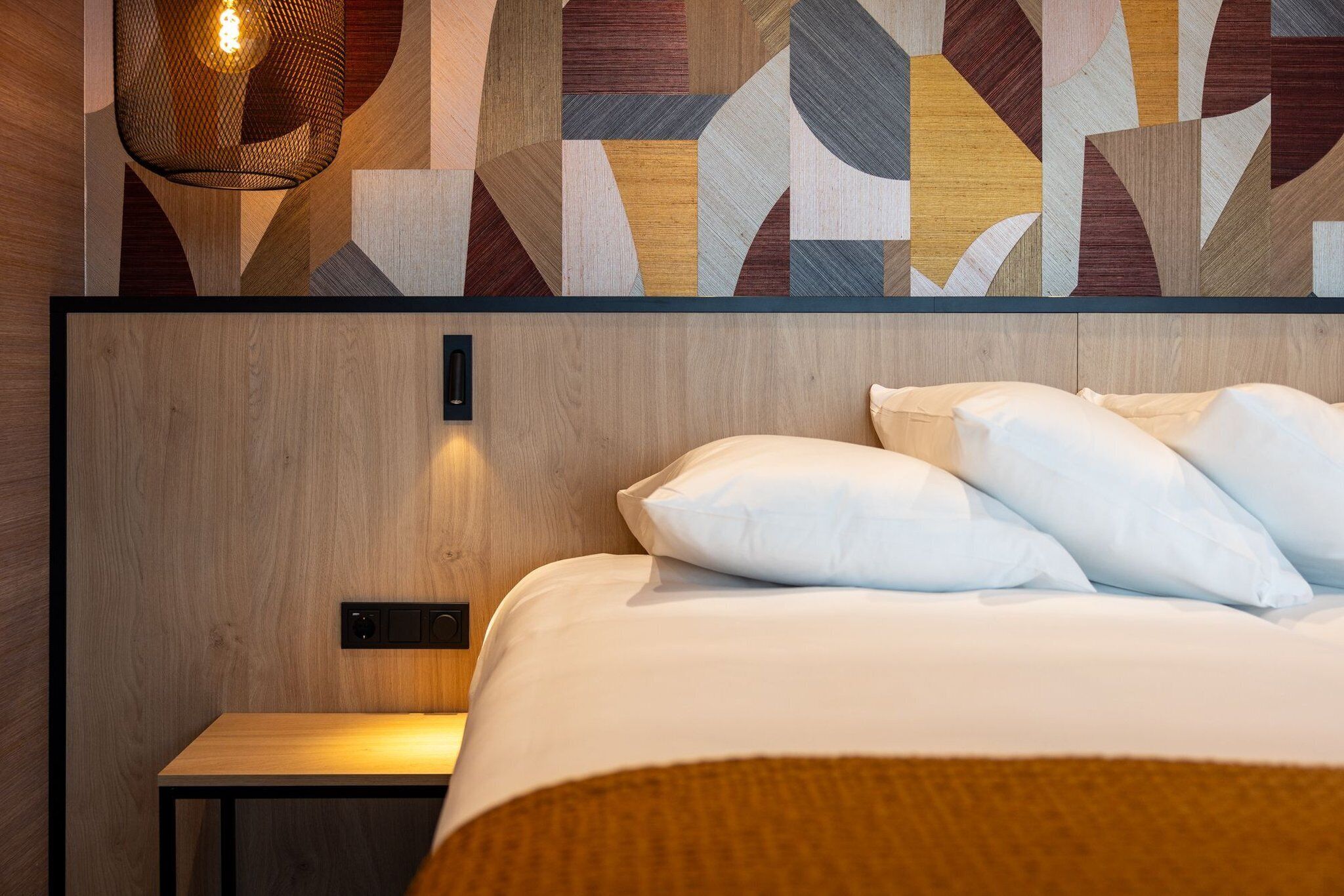 Quarto de hotel moderno com cabeceira de madeira, cama com roupa de cama branca e uma obra de arte geométrica na parede acima da cama.