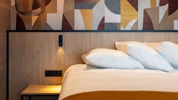 Quarto de hotel moderno com cabeceira de madeira, cama com roupa de cama branca e uma obra de arte geométrica na parede acima da cama.