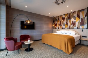 Une chambre d’hôtel avec un lit, une chaise rouge, une petite table, un téléviseur fixé au mur et une moquette à motifs.