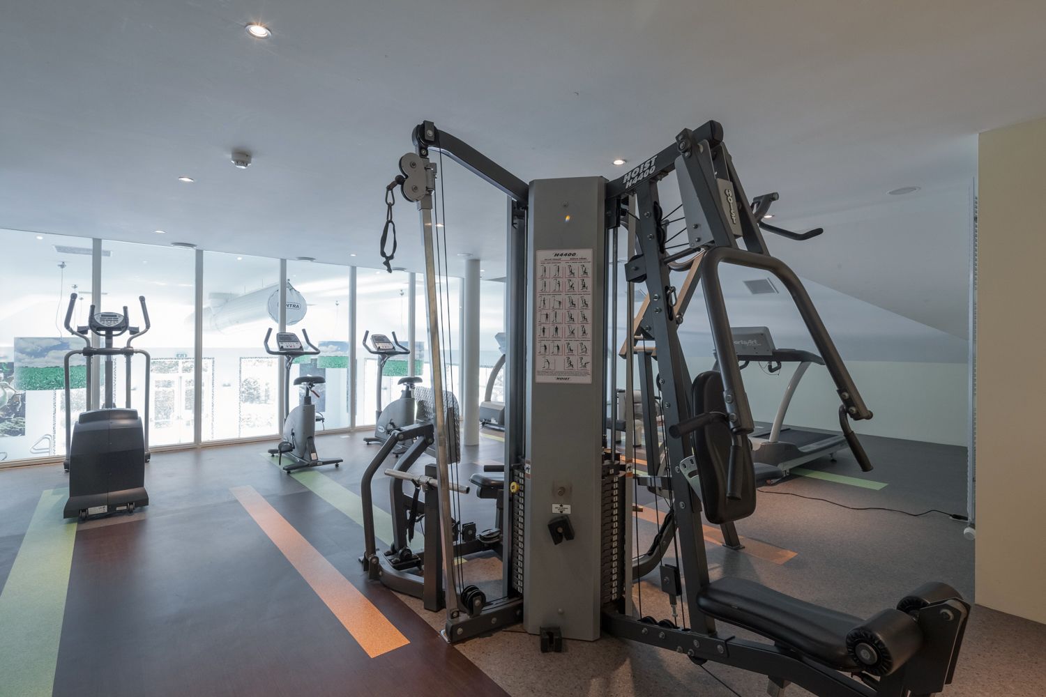 Un gimnasio moderno con máquinas de cardio, equipo de levantamiento de pesas y amplias ventanas con vista a la ciudad.