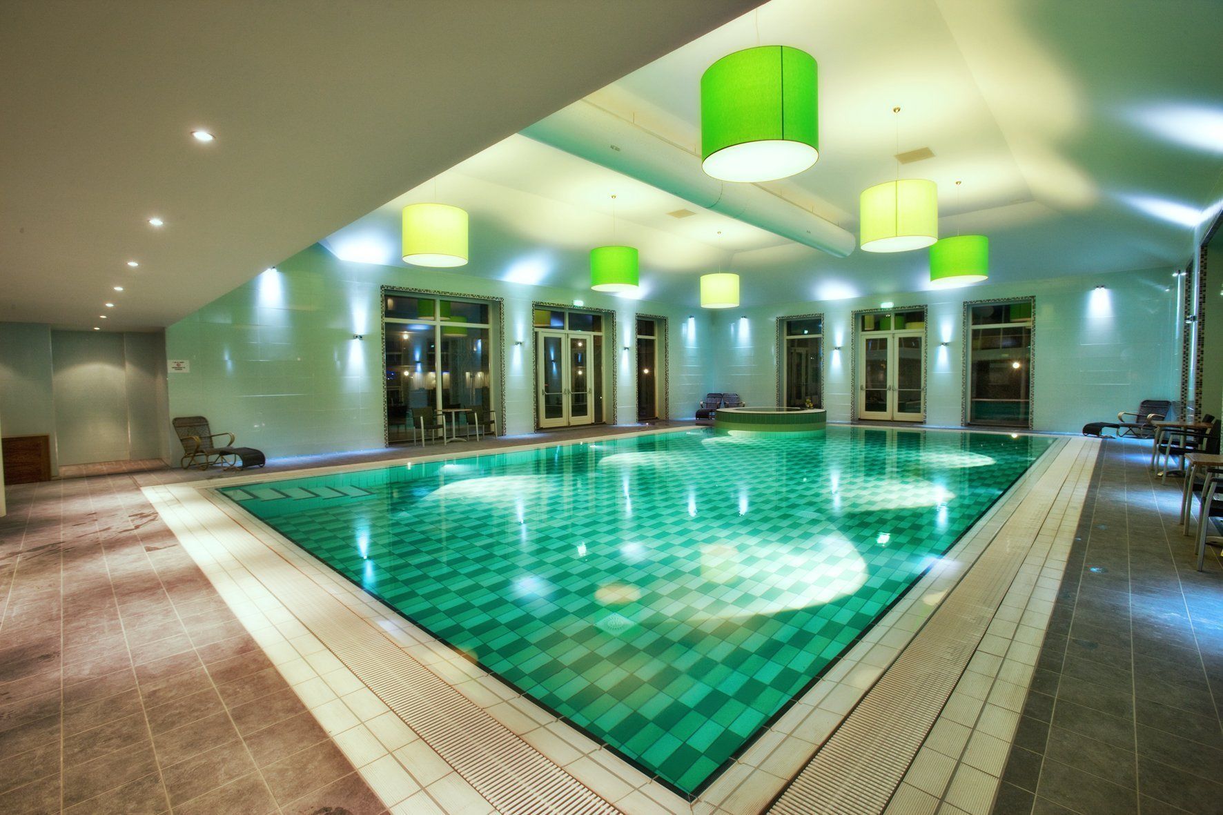 Uma grande piscina interior com pavimento xadrez, rodeada por cadeiras e mesas, e luminárias pendentes verdes.
