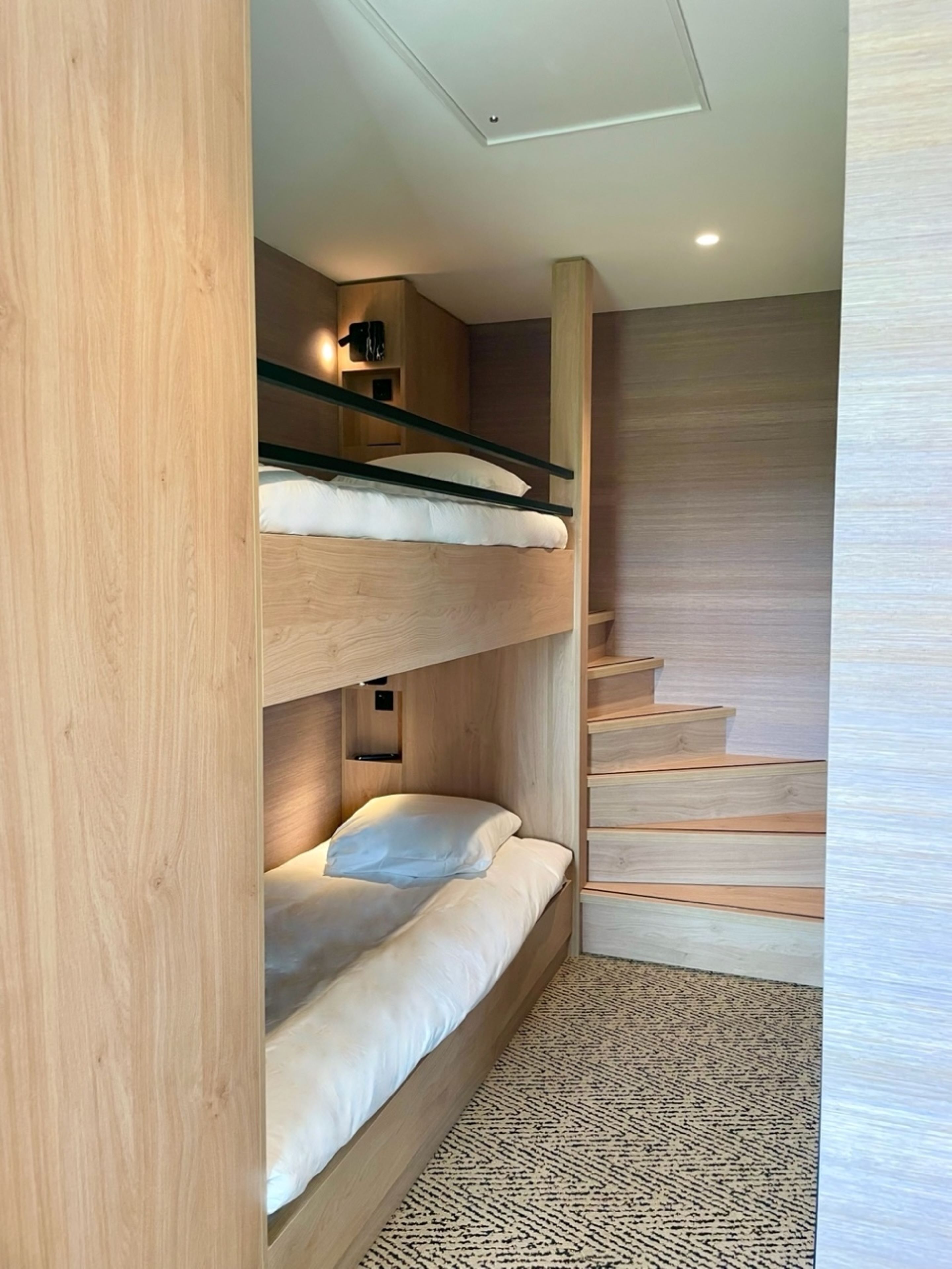 A bunk bed with a wooden finish, a small shelf, and a patterned rug.