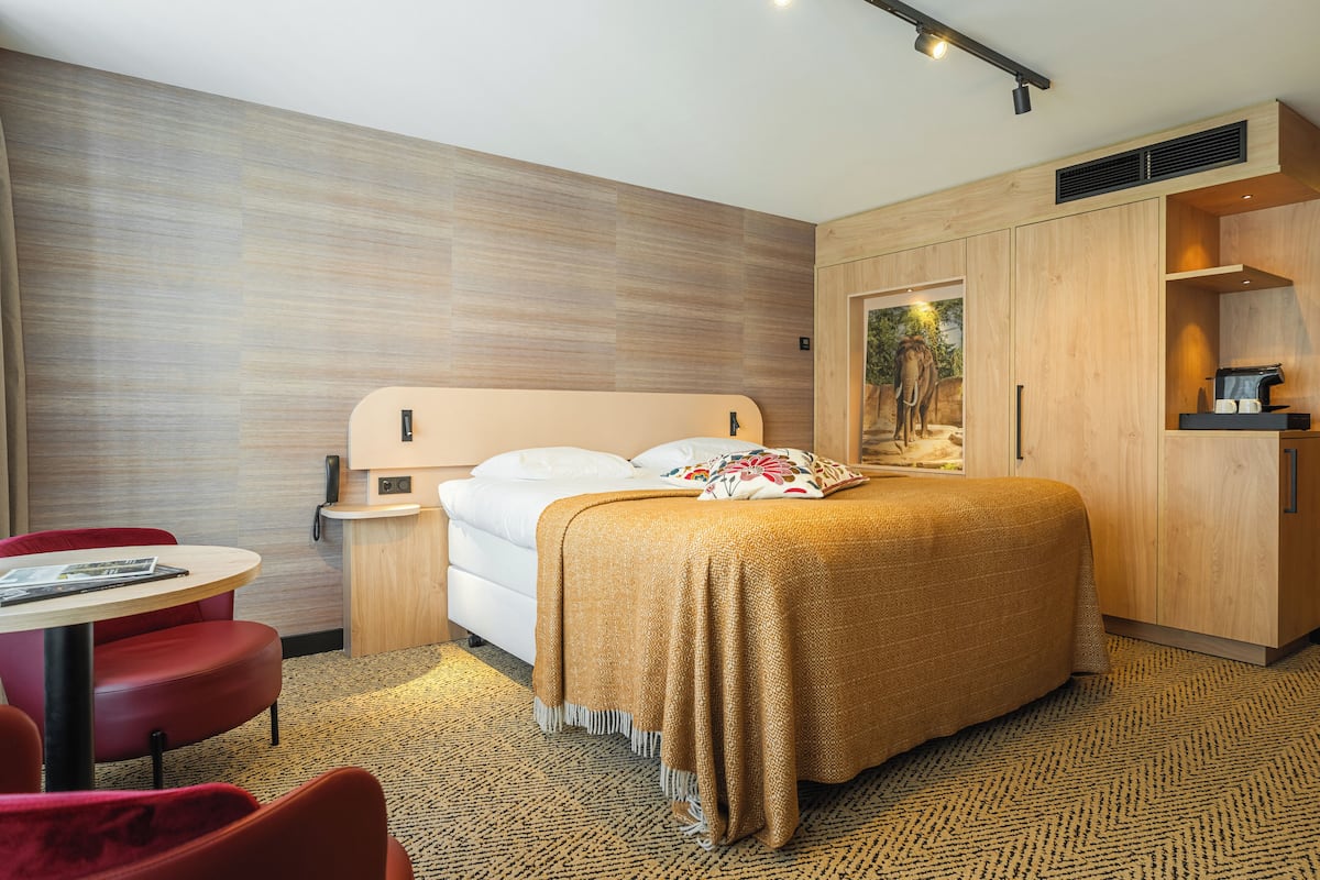 A modern hotel room with a bed, a desk, a chair, and a painting on the wall.