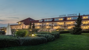 Un hotel con un letrero llamativo que dice "HOTEL EMMEN". Presenta un edificio de varios pisos con balcones y ventanas grandes. El hotel está rodeado de jardines bien cuidados y una fuente.