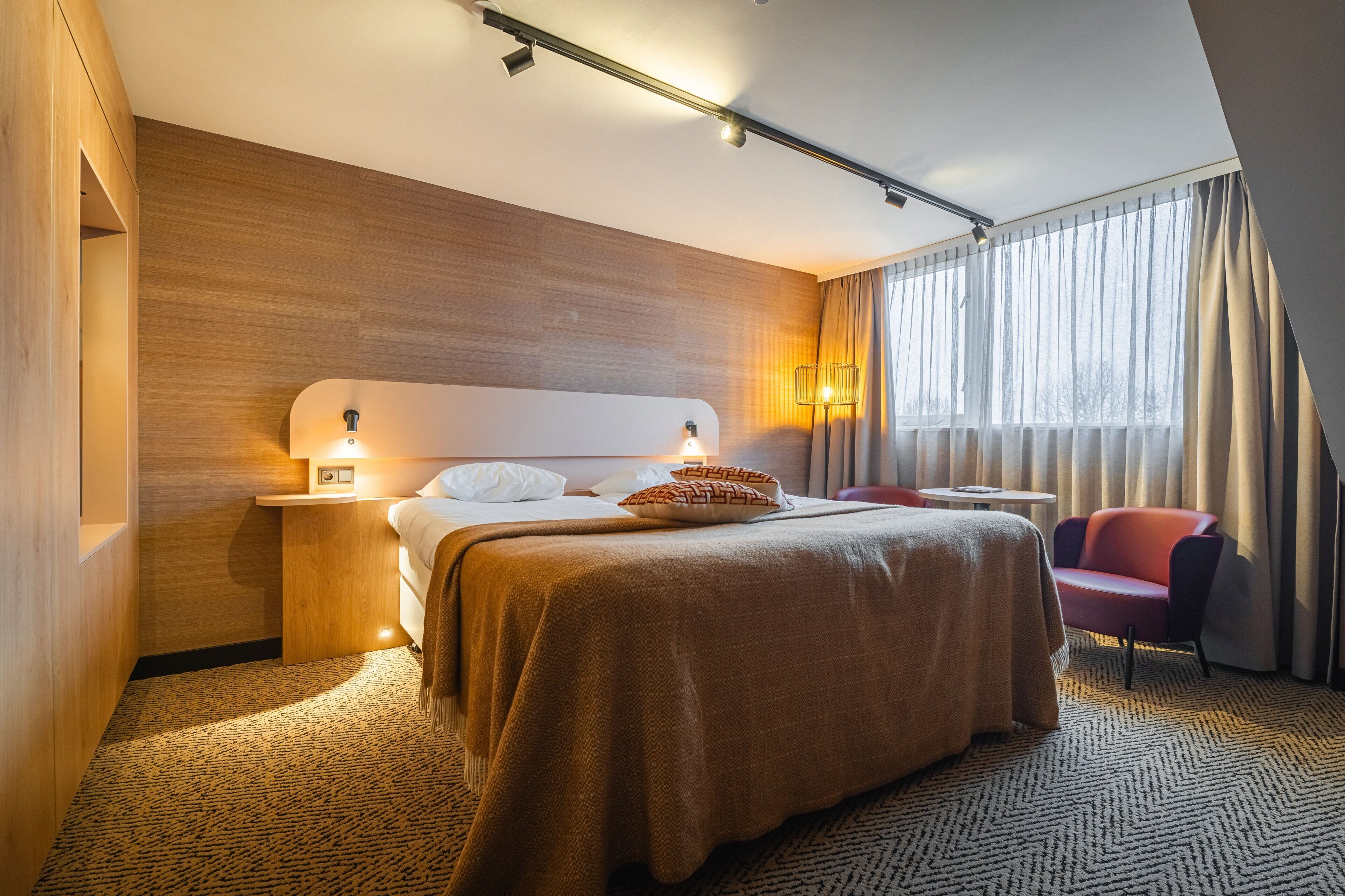 A hotel room with a large bed, a nightstand, a chair, a window with curtains, and a wooden wall.
