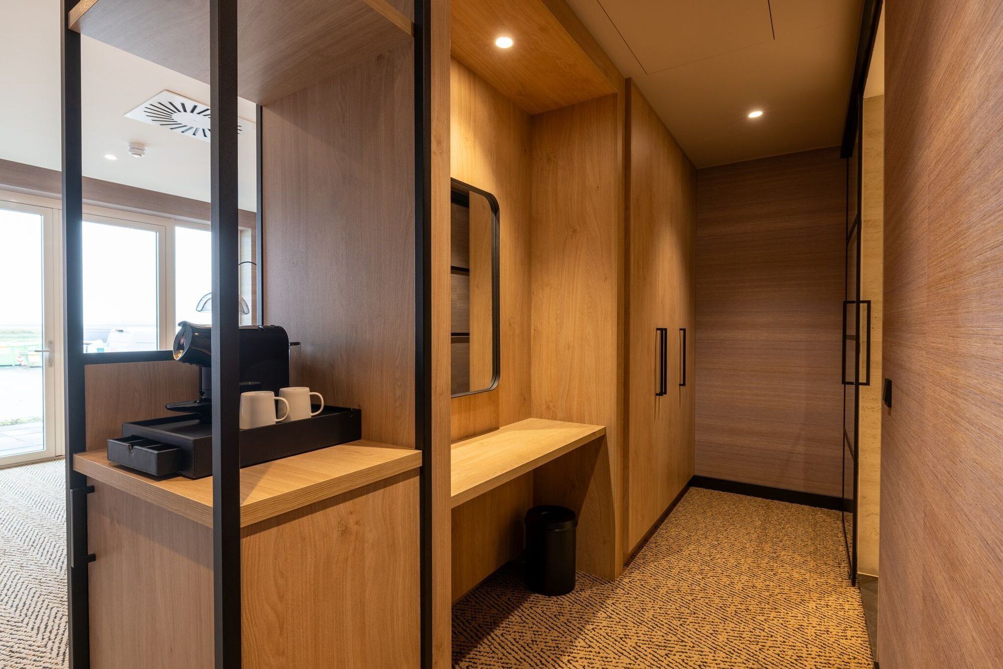 A modern hotel room with a wooden interior, a desk with a coffee machine, and a large mirror.