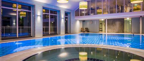 An indoor swimming pool with clear blue water, surrounded by walls with large windows and a modern ceiling design.