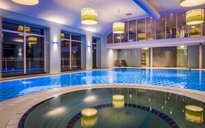 Une piscine intérieure à l’eau d’un bleu cristallin, entourée de murs dotés de grandes fenêtres et surmontée d’un plafond au design moderne.