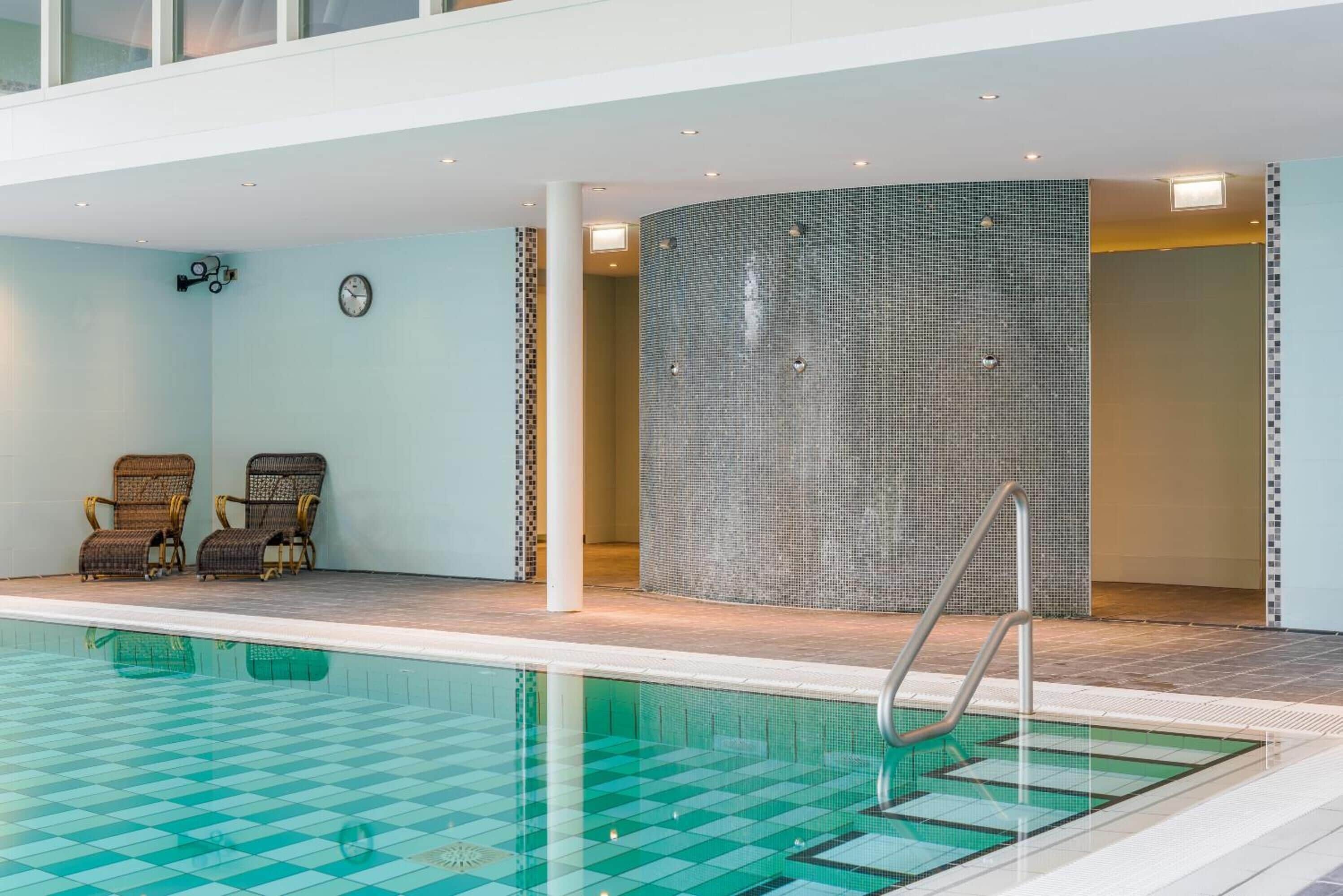 An indoor swimming pool with a checkered floor, two wicker chairs, a wall clock, and a tiled wall.