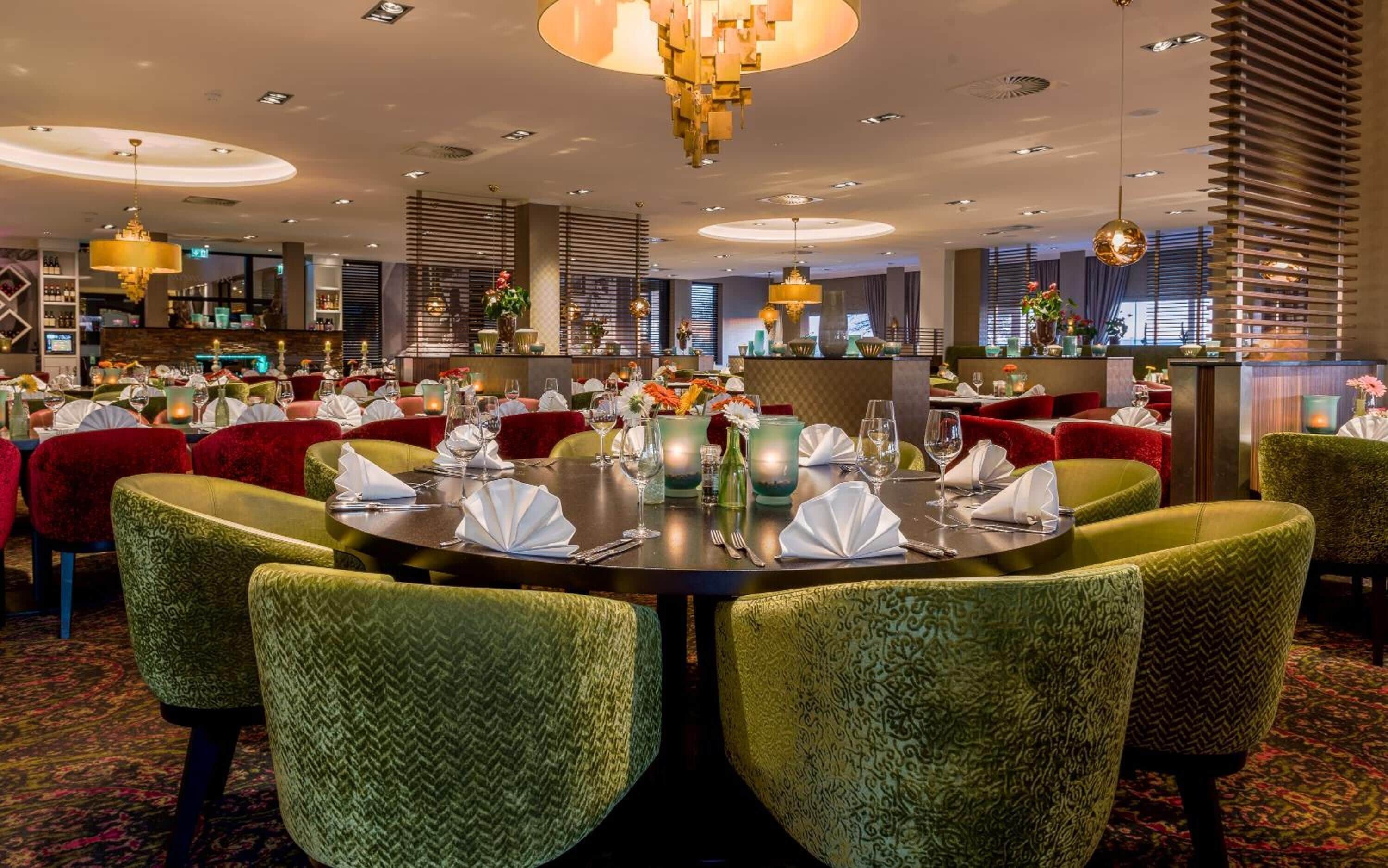 Una sala ristorante con tavoli rotondi apparecchiati, circondata da sedie imbottite verdi.