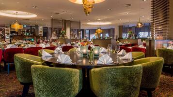 Una sala ristorante con tavoli rotondi apparecchiati, circondata da sedie imbottite verdi.