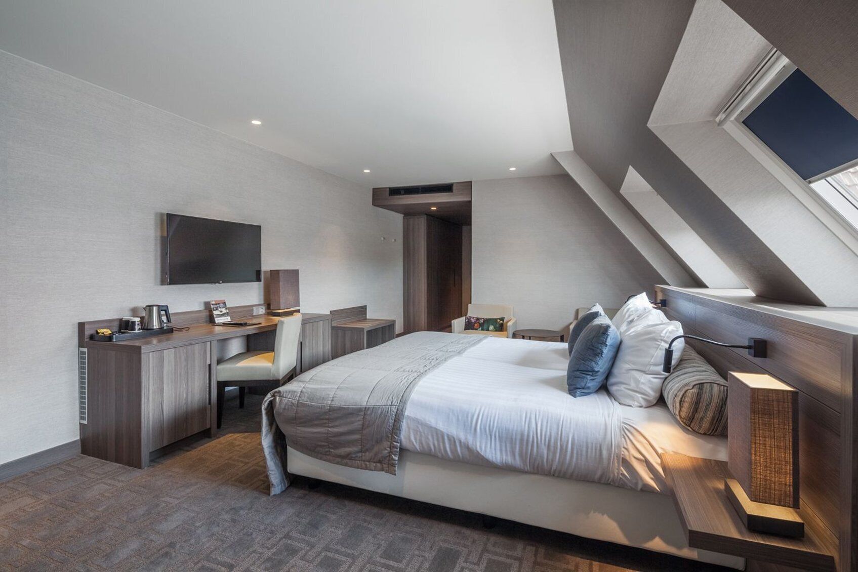 A modern bedroom with a bed, desk, and chair, a flat-screen TV, and a skylight.