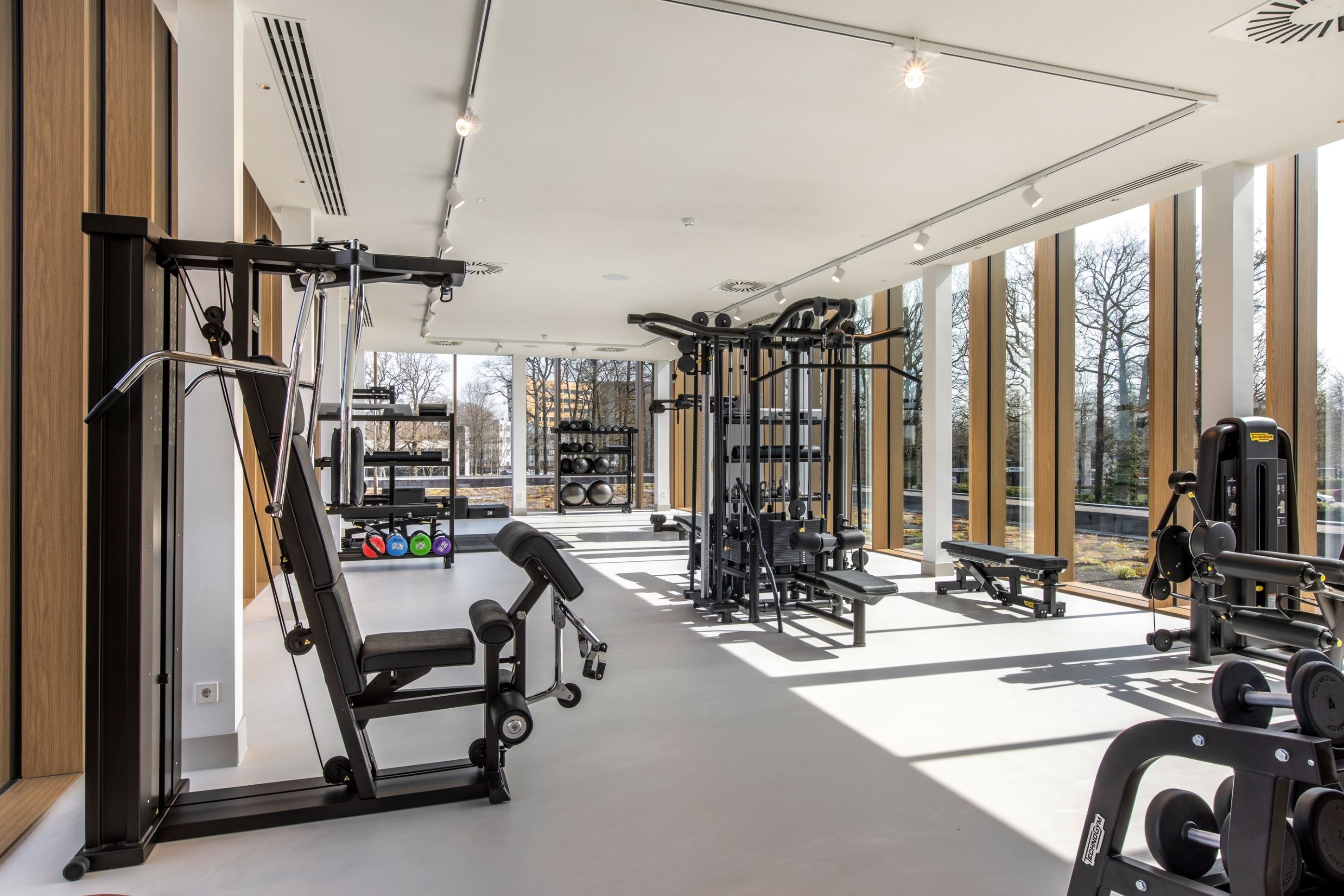 A modern gym with various exercise equipment, large windows, and a bench.
