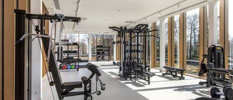 A modern gym with various exercise equipment, large windows, and a bench.
