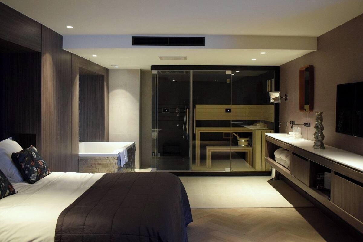 A modern bedroom with a large bed, a glass shower enclosure, a small desk, and a vanity with a mirror.