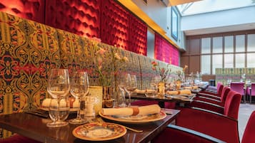 A restaurant with a long table set for dining, featuring a patterned wall and red upholstered chairs.