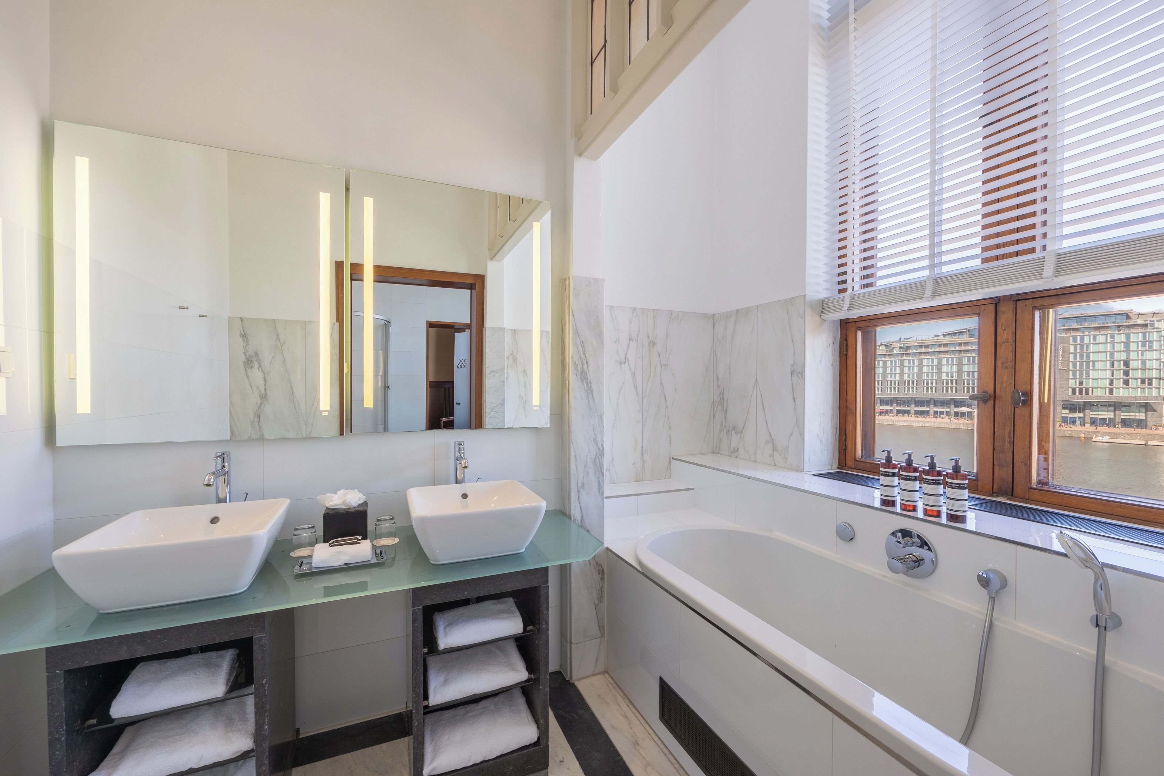 A modern bathroom with two sinks, a large bathtub, and a window with blinds.