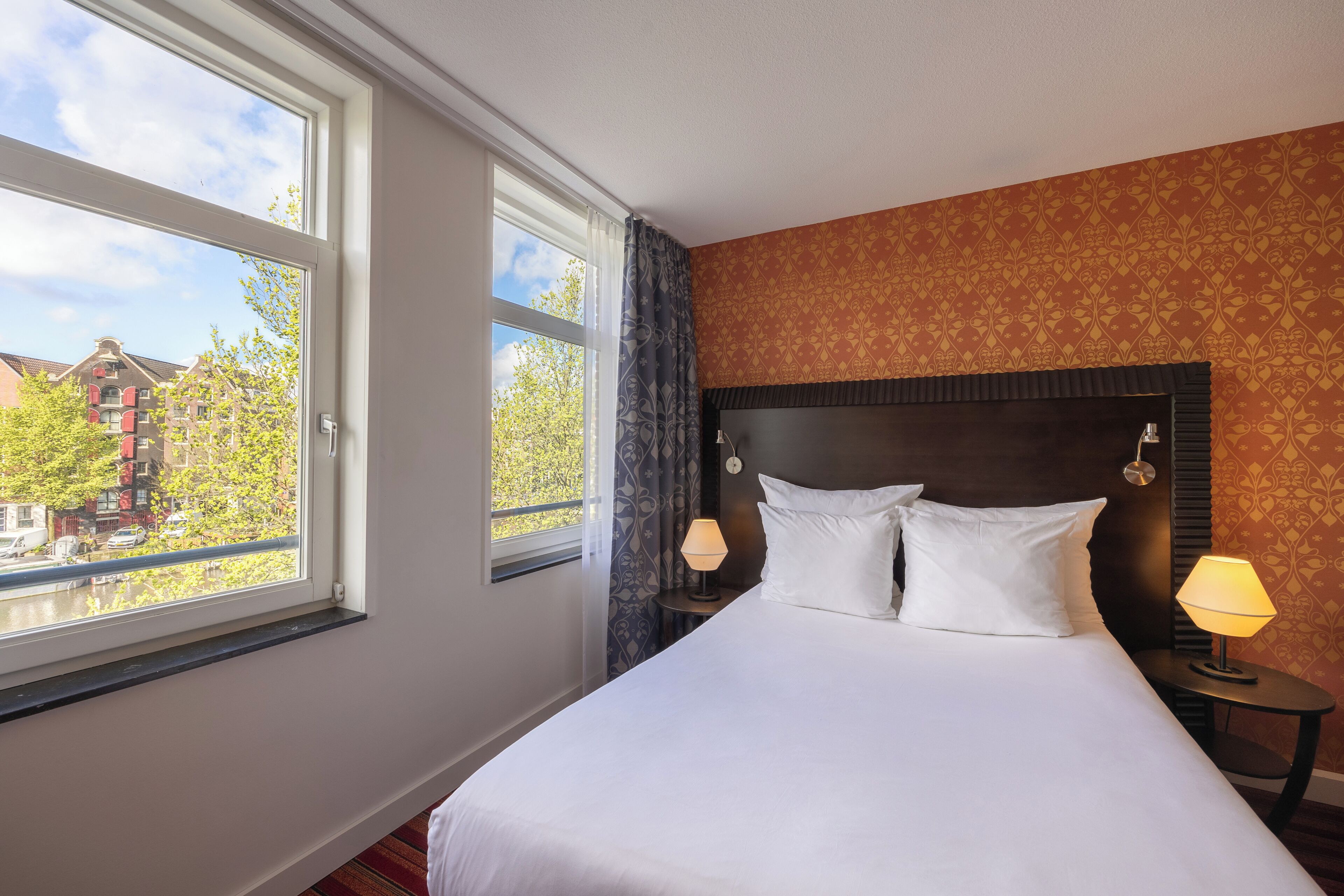 A hotel room with a large bed, two bedside lamps, a window with curtains, and a patterned wall.