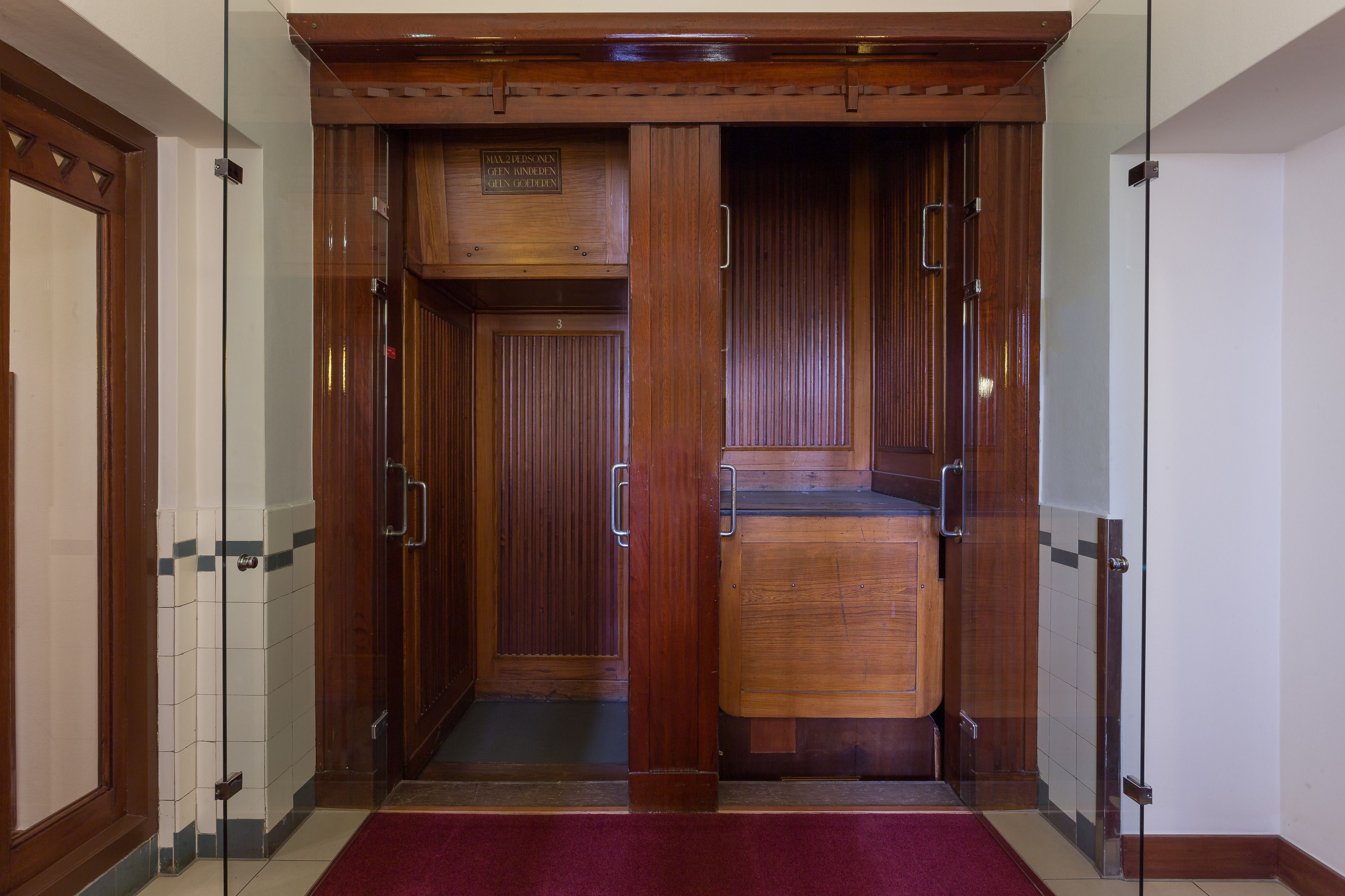 An elevator with wooden doors and a metal handle, leading to a room with a tiled floor and a red carpet.