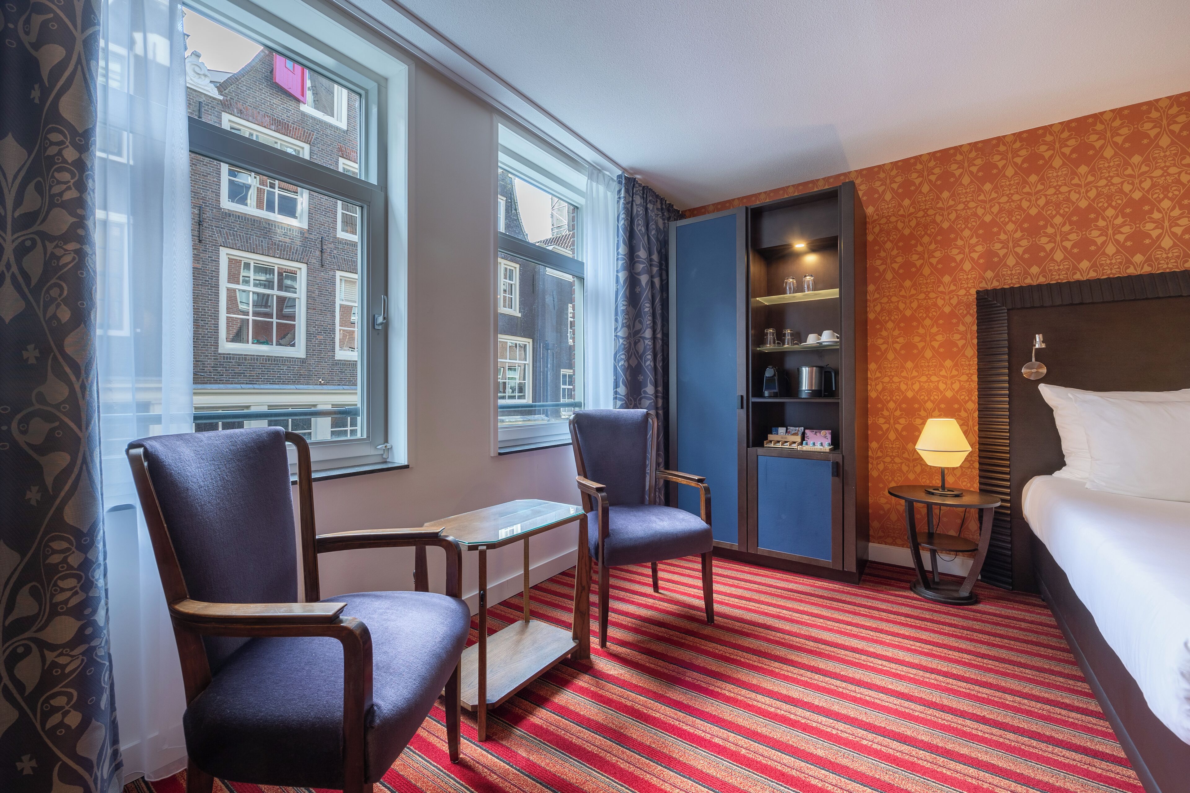 Une chambre d’hôtel avec un lit, deux fauteuils, une petite table, une lampe et une vue sur un immeuble à travers de grandes fenêtres.