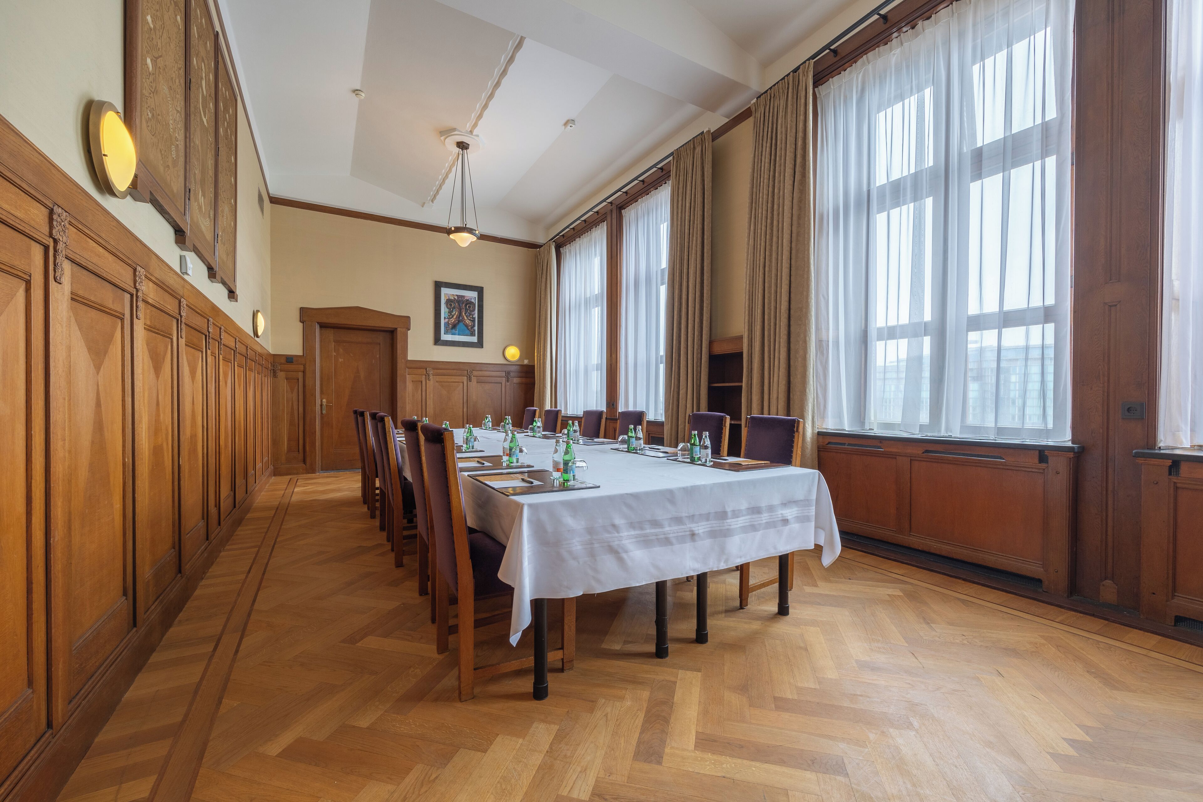 A long dining table with chairs, wooden paneling on the walls, and large windows with curtains.