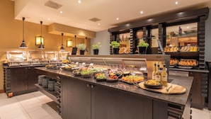 Uma área de buffet com diversas estações de comida, incluindo saladas, frutas e pão.