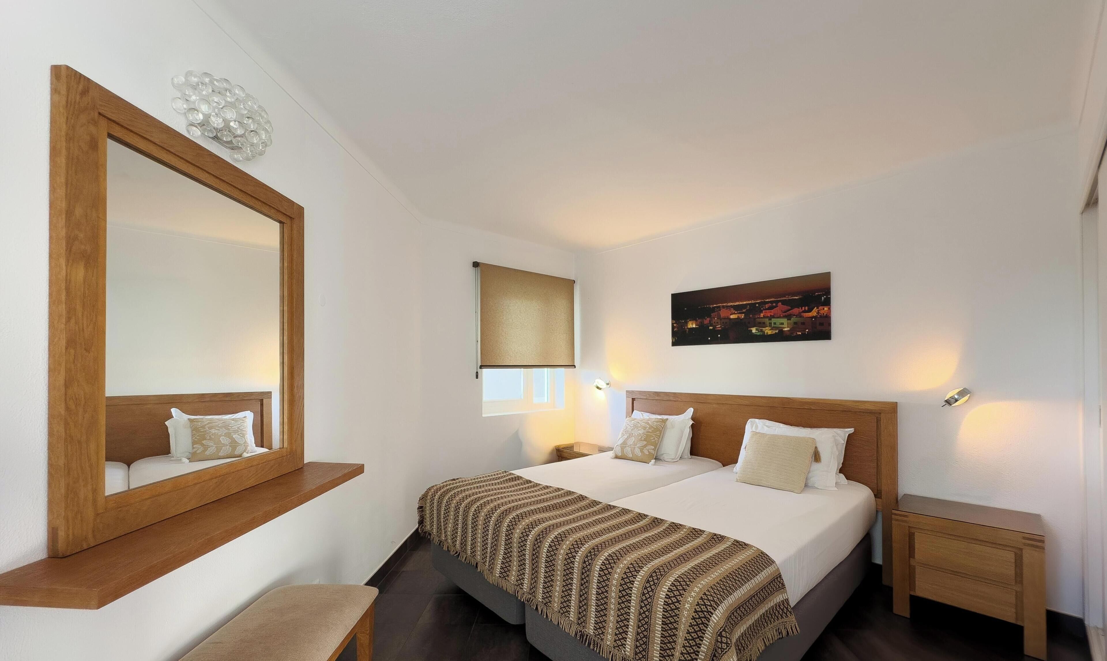 Een hotelkamer met een grote spiegel, een bed met gestreept beddengoed, een nachtkastje en een stadslandschap schilderij.