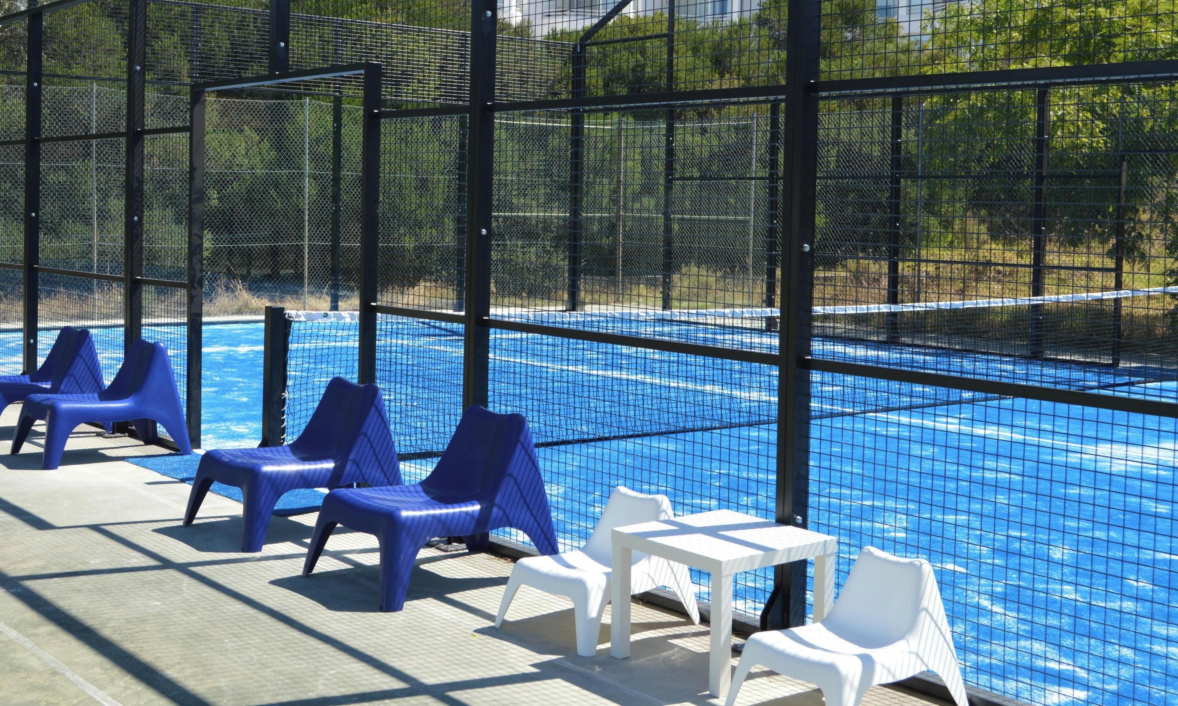 A swimming pool with blue water, surrounded by a black fence. There are blue and white lounge chairs and a small white table.