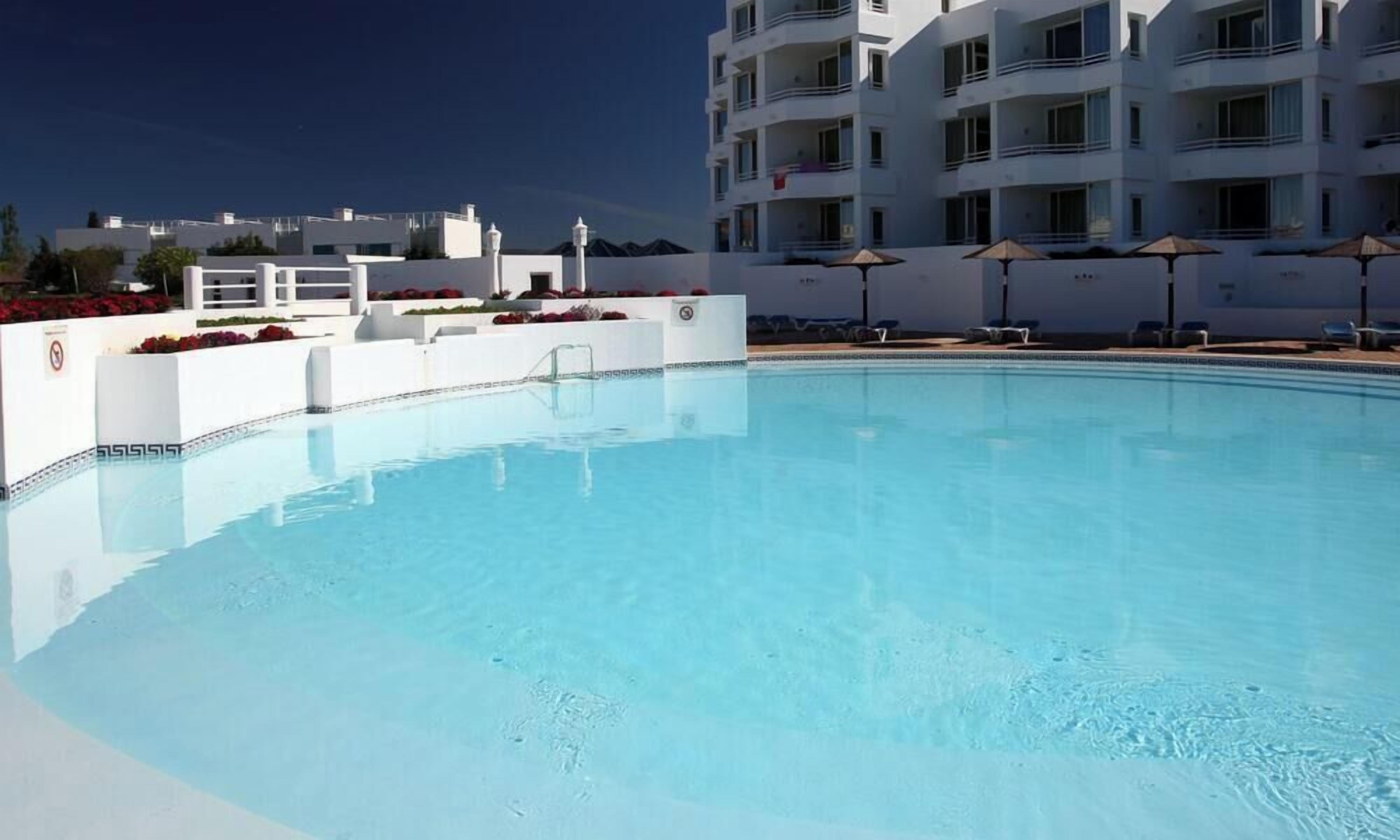 A swimming pool with lounge chairs and umbrellas, surrounded by a white building with balconies.