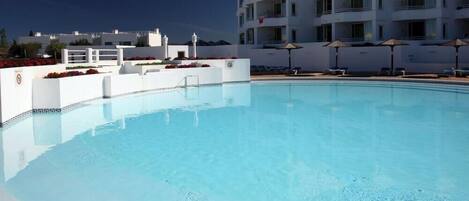 A swimming pool with lounge chairs and umbrellas, surrounded by a white building with balconies.