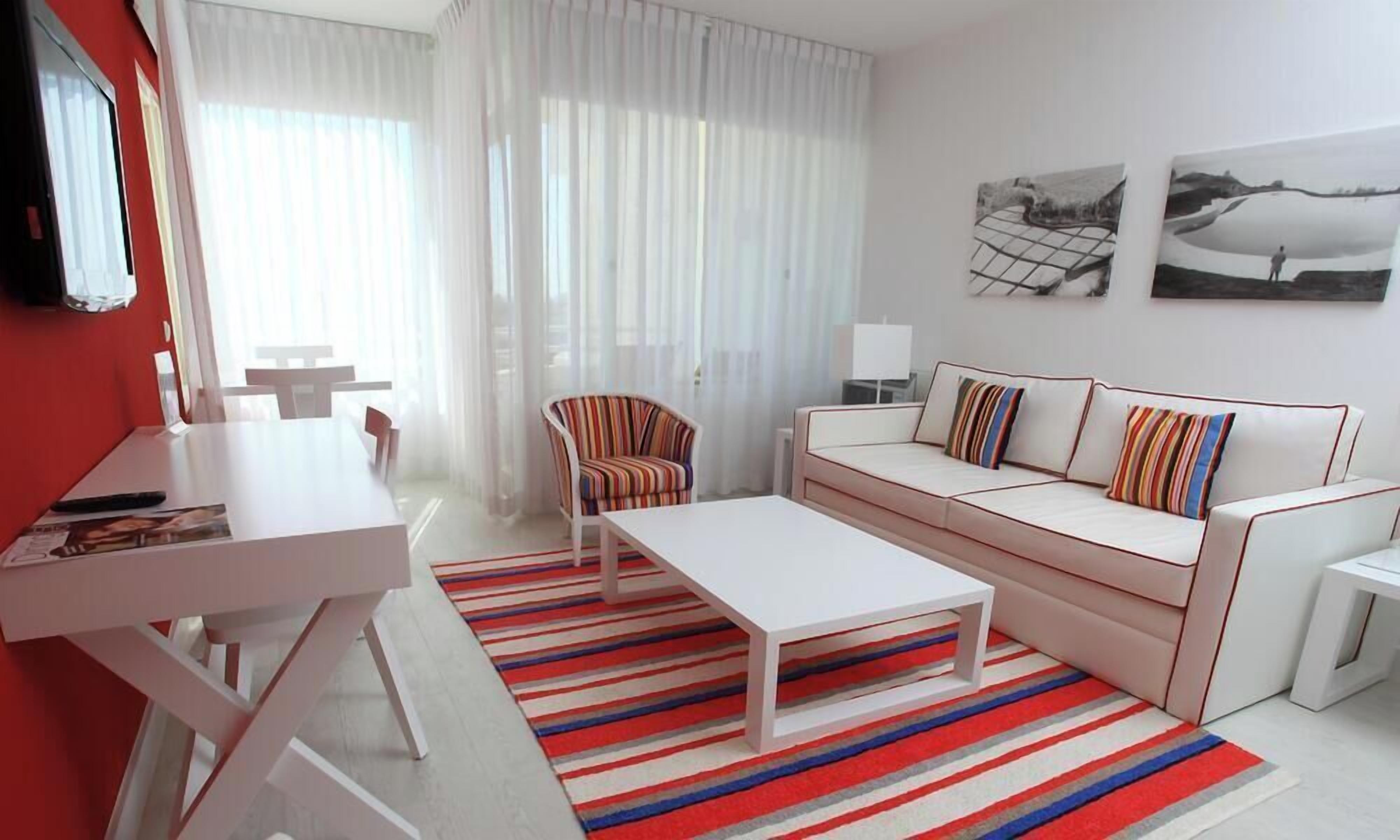 A modern living room with a white sofa, striped armchair, and a coffee table. There is a dining table with chairs, a TV mounted on the wall, and artwork on the walls.