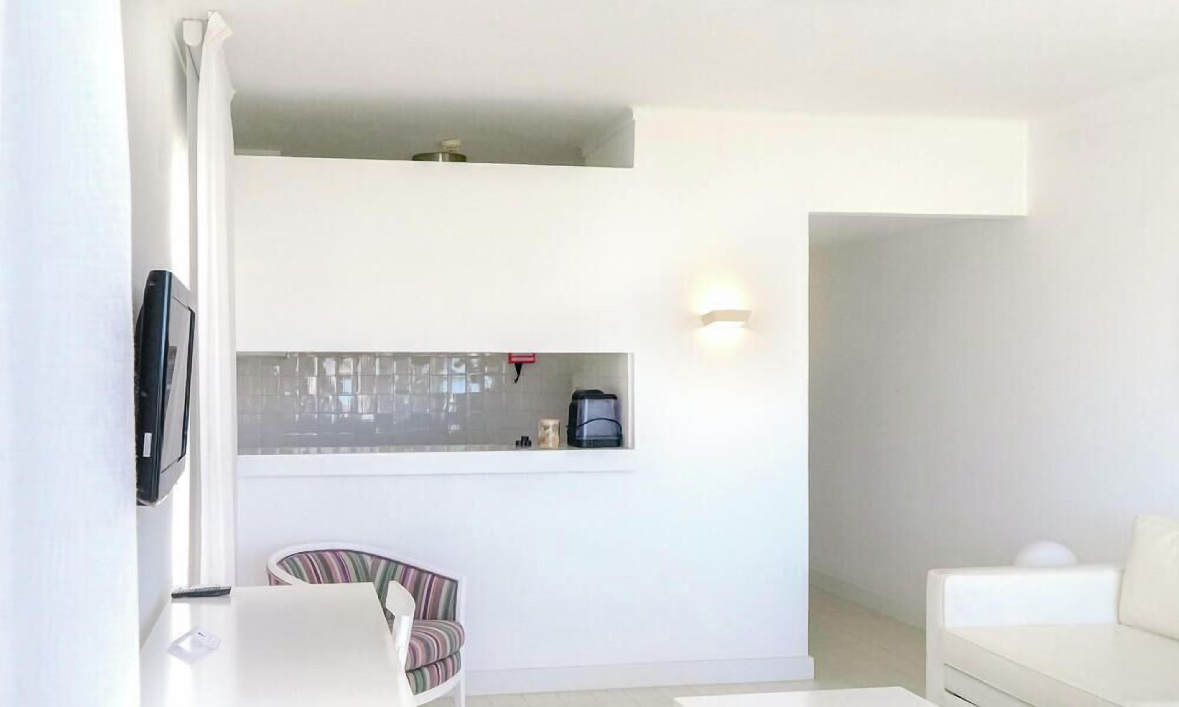 Un salon moderne et minimaliste, avec un canapé blanc, un fauteuil à rayures, un téléviseur à écran plat fixé au mur et un petit coin cuisine équipé d’une machine à café.