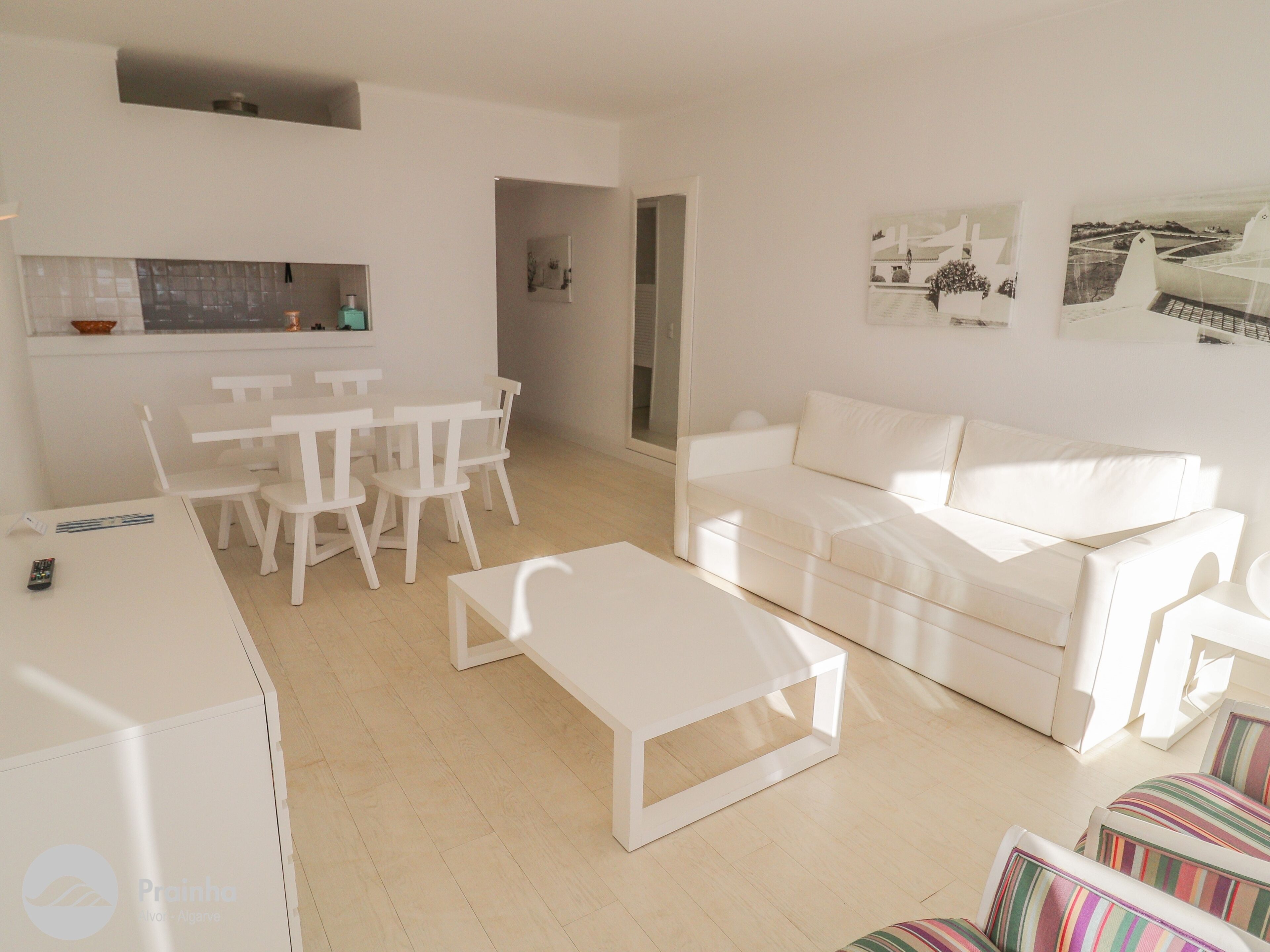 Un salon moderne avec un canapé blanc, une table à manger avec des chaises, une table basse et un coin cuisine équipé d’un réfrigérateur et d’un évier.
