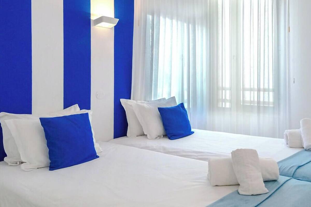 A hotel room with two beds, white bedding, blue pillows, and a large window with sheer curtains.