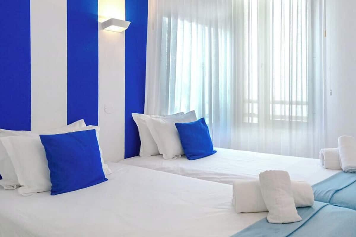 Une chambre dâhĂŽtel avec deux lits, du linge de lit blanc, des oreillers bleus et une grande fenĂȘtre avec des rideaux lĂ©gers.