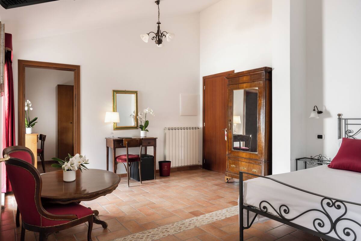 A bedroom with a bed, a desk with a chair, a table with chairs, a mirror, and a chandelier.