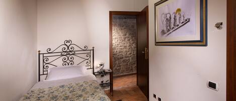 Un dormitorio con cama, mesita de noche, un cuadro enmarco, pared de piedra y puerta de madera.