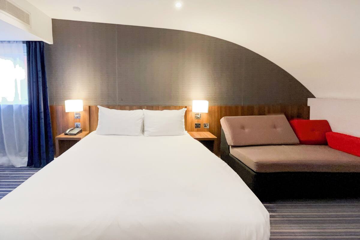 Habitación de hotel con una cama grande, dos mesitas de noche con lámparas, un sofá con cojines rojos y un diseño de pared curvo.