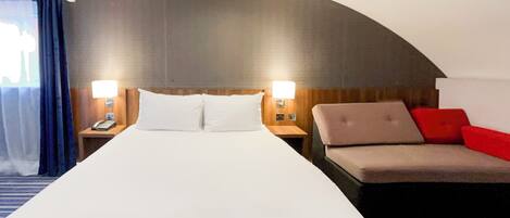 A hotel room with a large bed, two bedside tables with lamps, a sofa with red cushions, and a curved wall design.