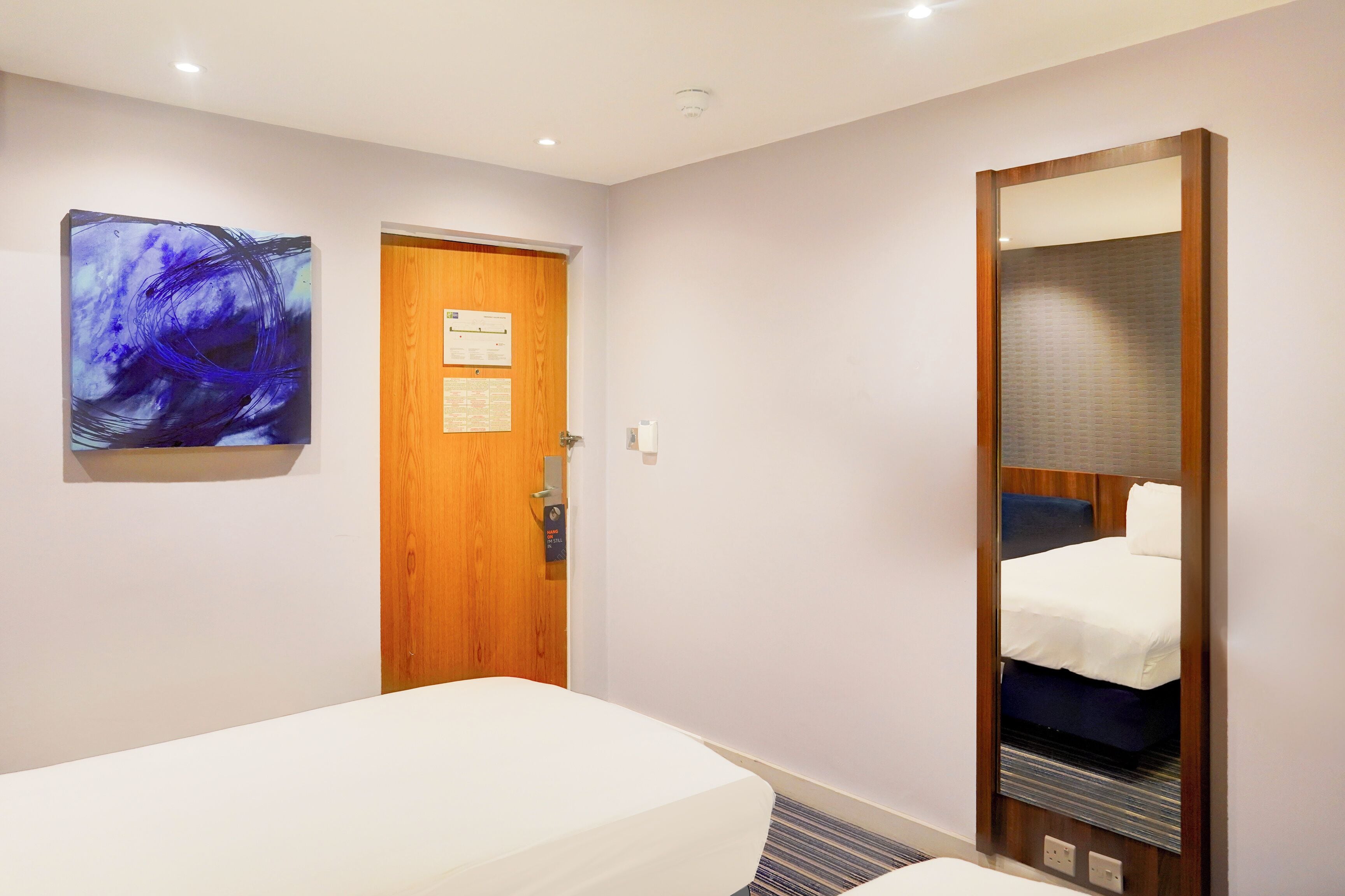 Habitación de hotel con una cama individual, una puerta de madera, un espejo grande y un cuadro abstracto colgado en la pared.