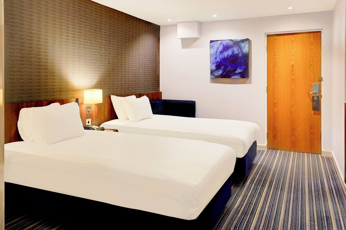 Habitación de hotel con dos camas, cabecero de madera, una obra de arte colgada en la pared y una puerta con cerradura.