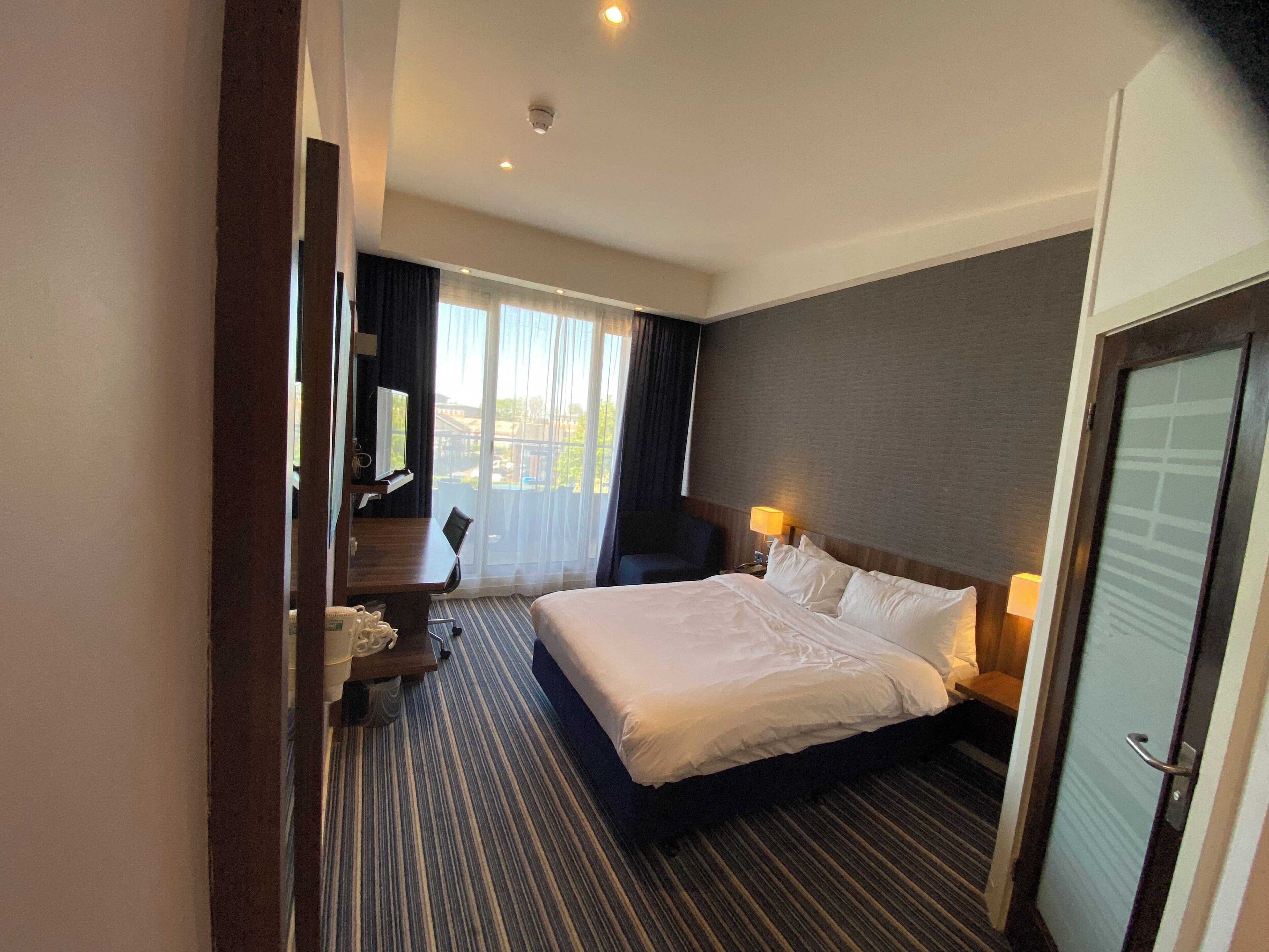 Habitación de hotel con cama, escritorio, silla, lámpara y vistas al exterior.
