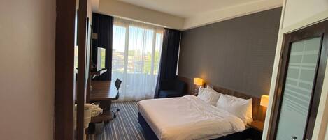Habitación de hotel con cama, escritorio, silla, lámpara y vistas al exterior.