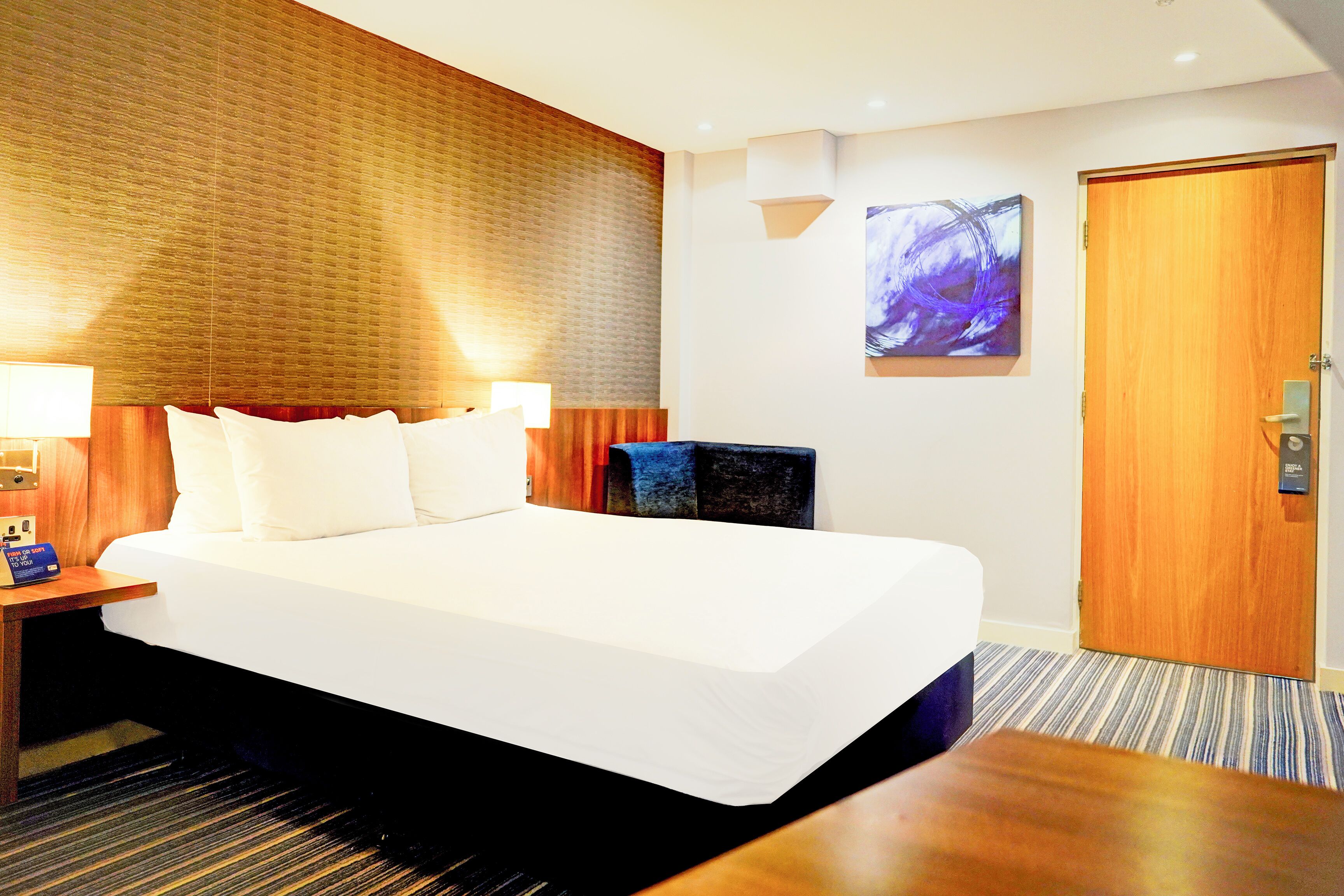 A hotel room with a large bed, a nightstand with a lamp, a wooden headboard, a painting on the wall, and a door.