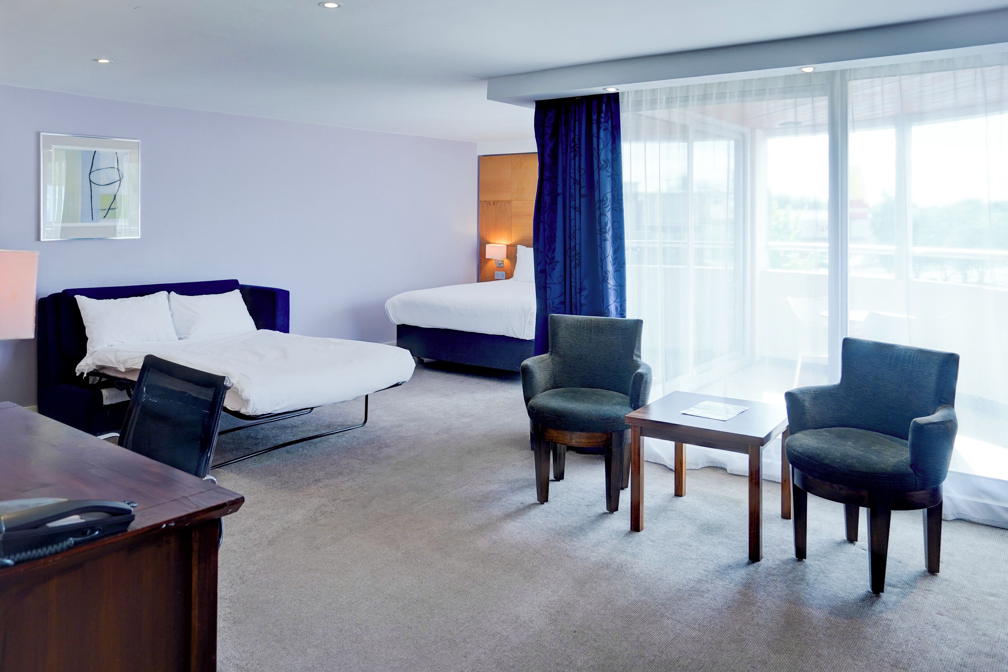 Een hotelkamer met een bed, twee fauteuils, een bureau en een groot raam met gordijnen.
